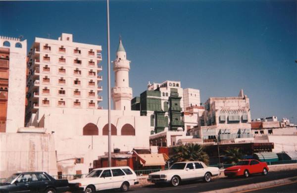 White minaret among Jeddah buildings, cars on the street under blue sky
