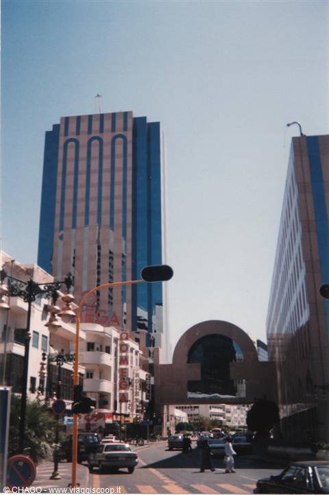 Mirror-glass blue skyscrapers above a busy street with cars and pedestrians in sunlight