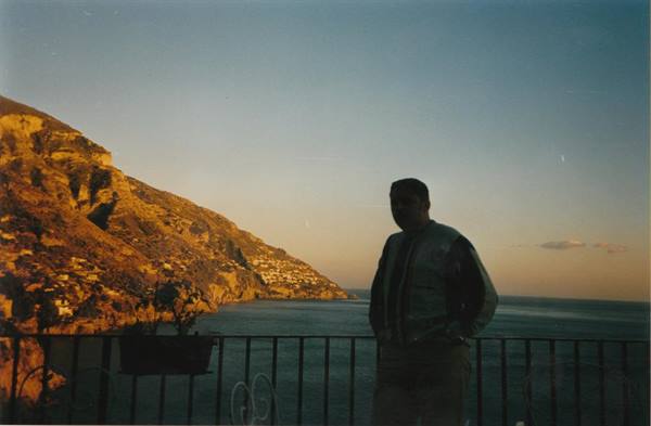Man in jacket by a railing, rocky coast with houses above the sea