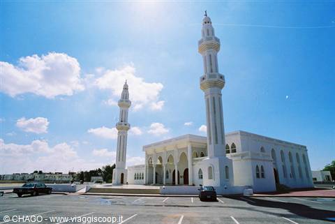 White mosque with two minarets, cars parked under blue sky with clouds