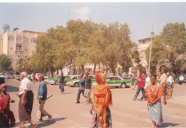 Pedestrians crossing a tree-lined square with green taxis and buildings