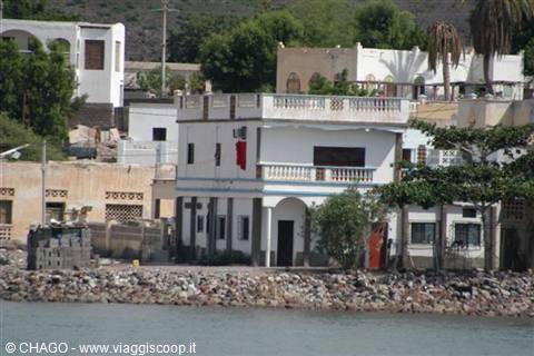 White building with balconies by rocky seashore, tropical houses nearby