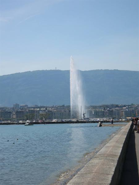 Water jet over Lake Geneva, stone promenade, boats and mountains behind