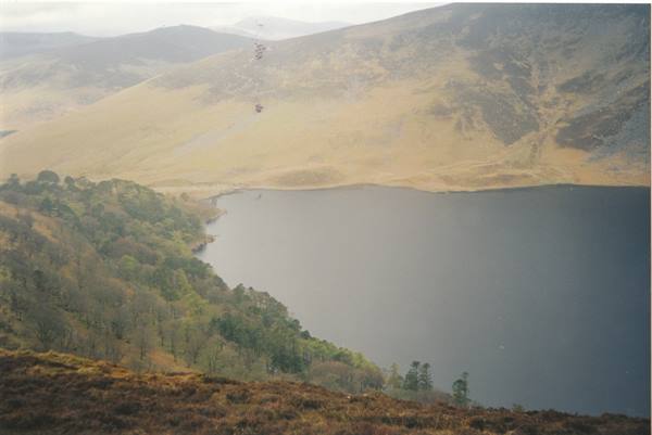 Grey lake surrounded by green woods and bare hills under a misty sky