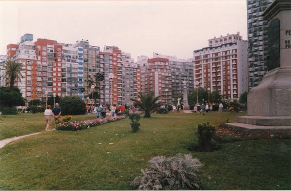 People strolling among flowerbeds in an urban square, colorful apartment buildings behind