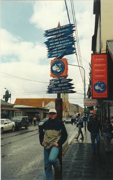 Tourist beneath Ushuaia signpost showing distances to world cities