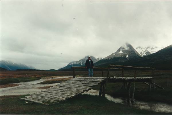 Man standing on a wooden log bridge, snowy mountains veiled by clouds