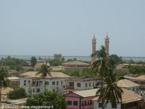 Slim minaret among low houses and palm trees, water on the horizon Banjul