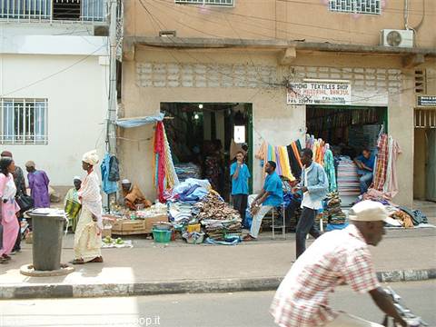Men sitting outside a fabric shop, pedestrians and a cyclist on the street