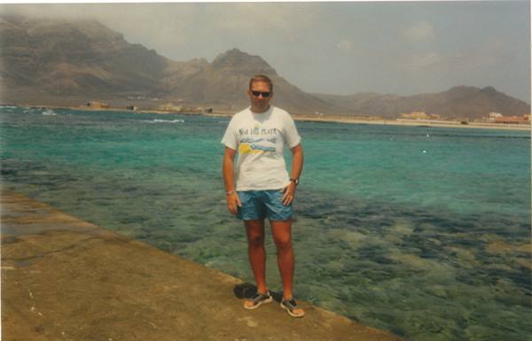 Man in white t-shirt and shorts on concrete pier facing turquoise sea and mountains