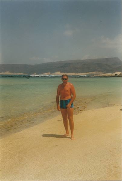 Man in blue swimsuit standing on light sand, turquoise sea and mountains behind