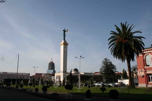 Column with golden statue in tree-lined square, tall palm and buildings