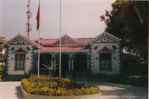 White and turquoise building with red roof, circular flowerbed and flag in front