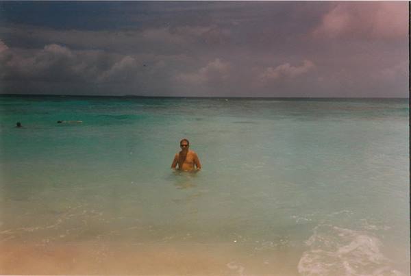 Man in swim trunks standing in turquoise water, pale sand beach, cloudy sky