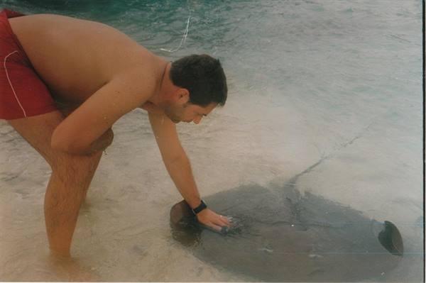Man in red swimsuit strokes a stingray in shallow sandy water