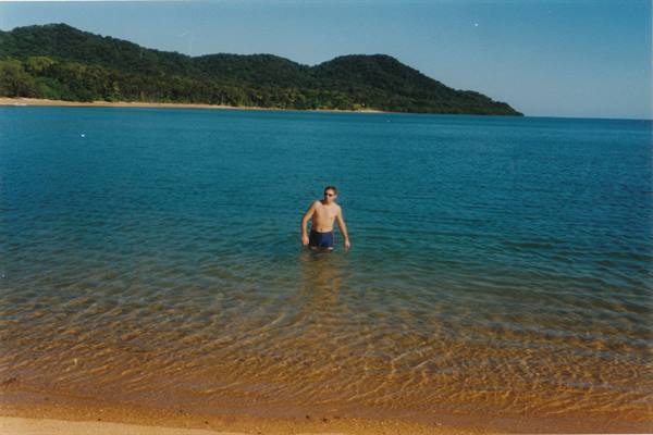 Man in swim trunks wading in turquoise water in front of green hills