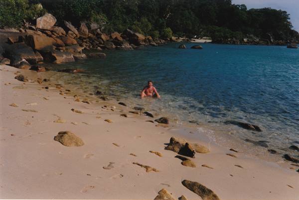 man in clear water among rocks and sand, vegetation along the shore