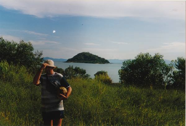 Man in hat among tall grass, tree-covered island in the sea