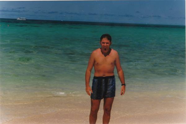 Man in dark swim shorts on the beach, turquoise sea with white ferry behind