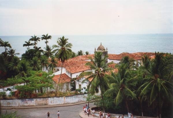Colonial church with terracotta roofs among palm trees, people walking by the sea