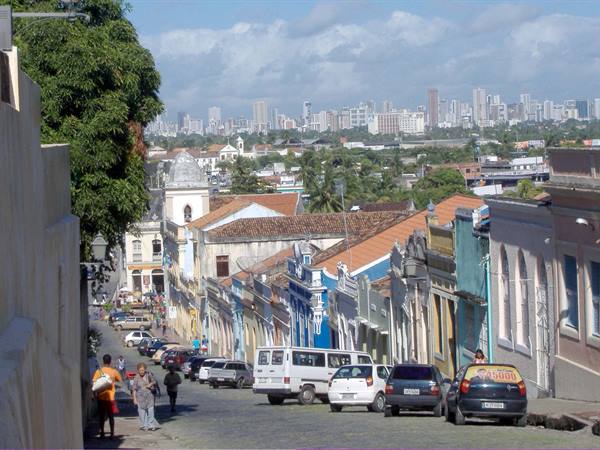 Steep street with colorful colonial houses and skyscrapers behind, Olinda