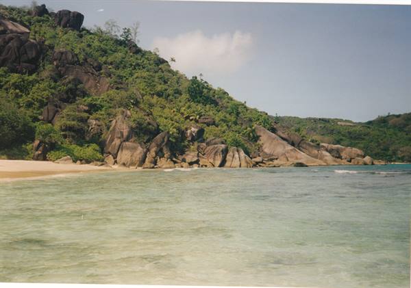 Clear turquoise water, sandy beach and granite boulders covered by green vegetation