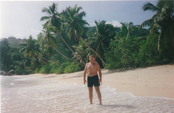 Man in black shorts standing in shallow water among palm trees and white sand