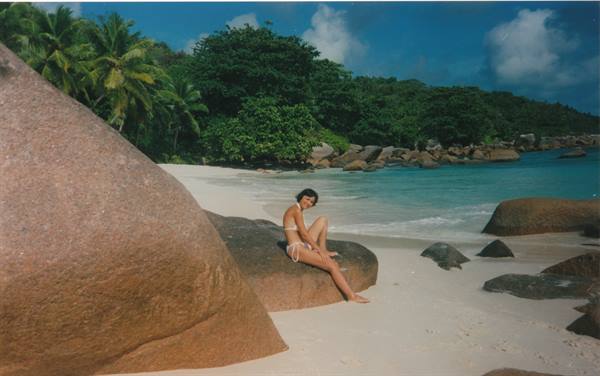 Woman in swimsuit sitting on large granite boulder, white sand, palms and turquoise sea
