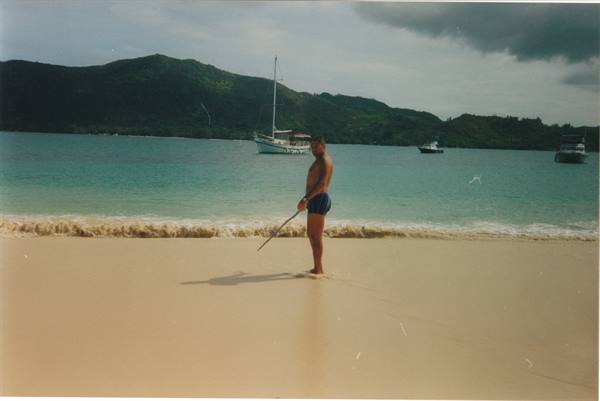 Man in swim trunks holding stick on beach, sailboat and green hills behind