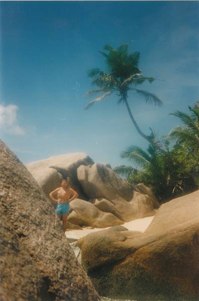 Man in swim trunks among giant granite boulders and leaning palm, Anse Source d'Argent