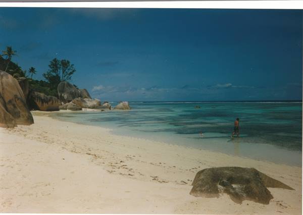 Man wading in shallow water among granite boulders and white sand, Anse Source d'Argent