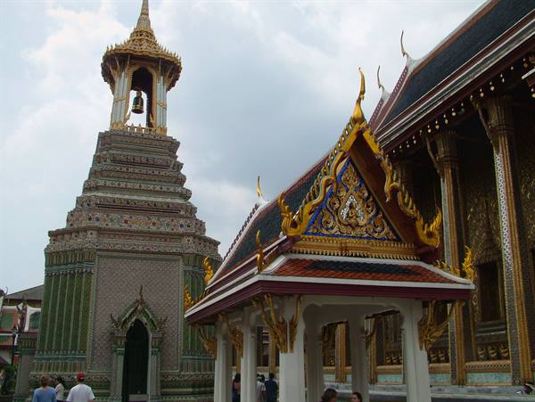 Gilded pavilion and tiled bell tower at Wat Phra Kaew, tourists under grey sky
