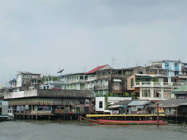 Stilt houses along the river, red longtail boat moored, grey sky