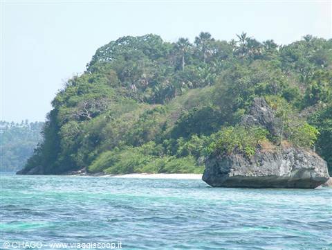 Large rock outcrop before white sand beach and green tropical hill