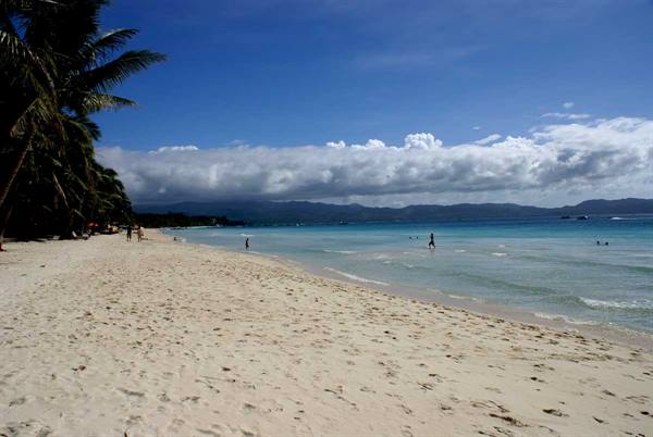 White sand beach with palm trees and swimmers in turquoise water, clouds on horizon