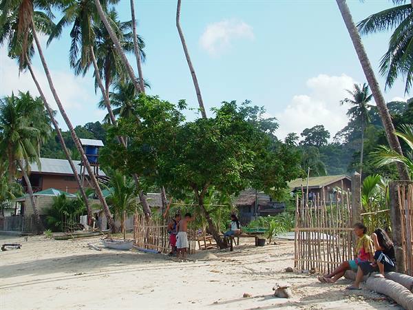 Sandy beach with leaning palm trees, wooden houses and children sitting near the bamboo fence