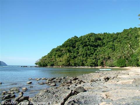 Sandy beach with dark rocks, lush green tropical hill and boat on turquoise sea