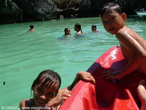 Smiling children in turquoise water next to a red kayak by cliffs