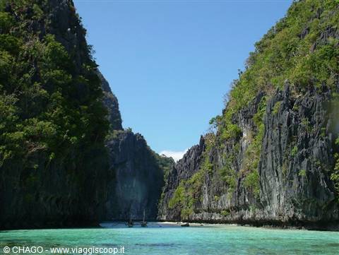 Turquoise lagoon between green limestone cliffs, fishing boats in the distance
