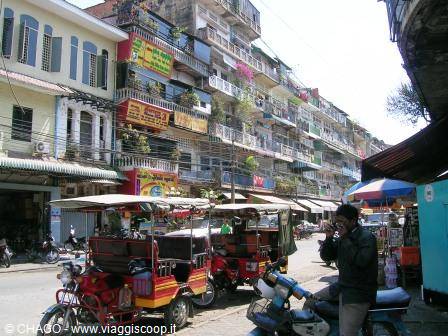 Tuk-tuks lined up under balconied buildings, colorful signs and people passing by