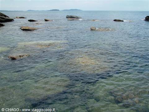 Granite rocks emerge from clear emerald water under a bright sky