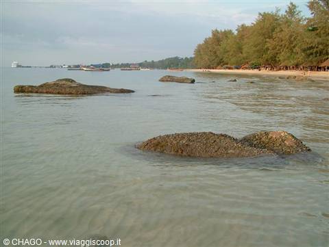 Grey rocks emerge from shallow water before a tree-lined beach and boats