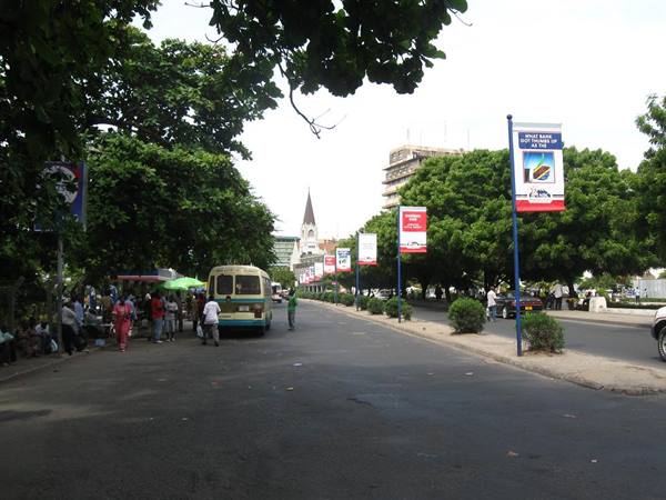 Tree-lined Lagos street with parked minibus, vendors and advertising banners