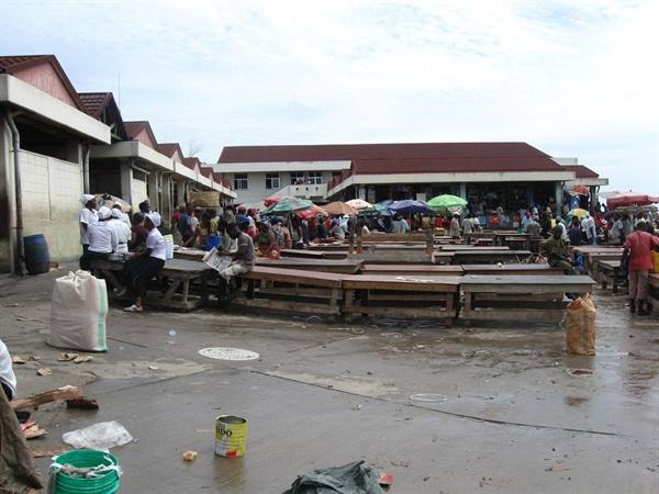 Open-air market with wooden stalls, colorful umbrellas and people under red-roofed pavilions
