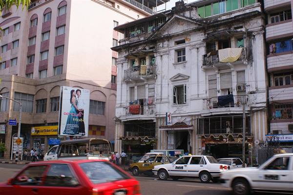 Taxis and cars passing weathered colonial building, busy traffic, Yangon