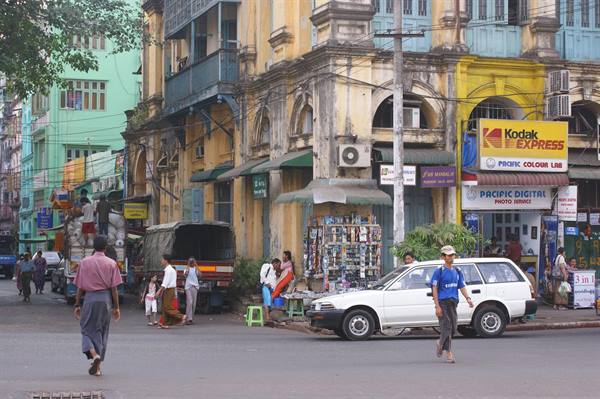 Men in longyi cross a downtown Yangon street past colonial buildings and Kodak signs