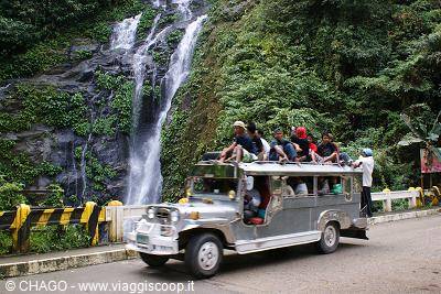 Tourists riding on top of a silver jeepney in front of a forest waterfall