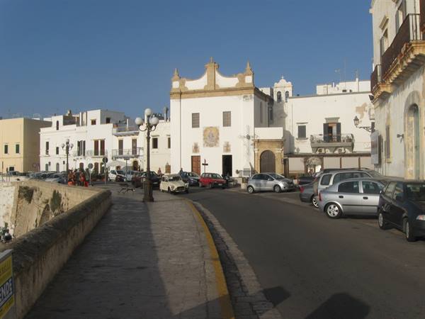 Square with white church and historic houses, parked cars under blue sky, Gallipoli