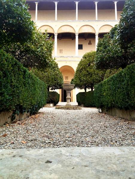 Stone fountain at the center of a gravel path, hedges and arched loggia