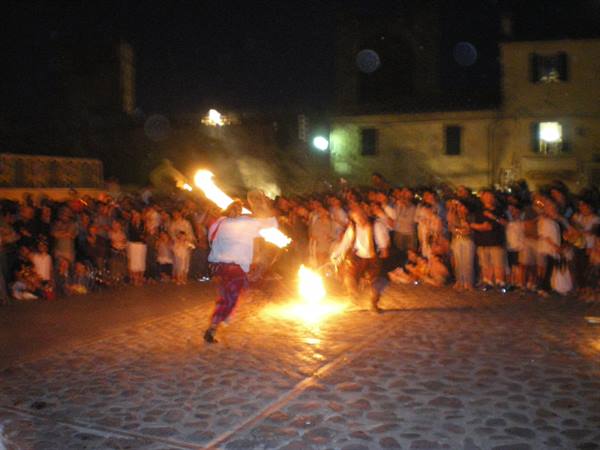 Night fire performance on cobblestones, crowd gathered among old buildings